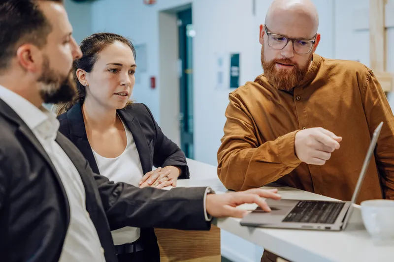 Three people in a business meeting discussing something on a laptop screen. One person is pointing at the screen while the others listen attentively.
