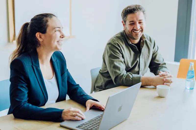 Two people sitting at a table, smiling and engaged in conversation. One person is using a laptop, and there is a cup and a bottle of water on the table.